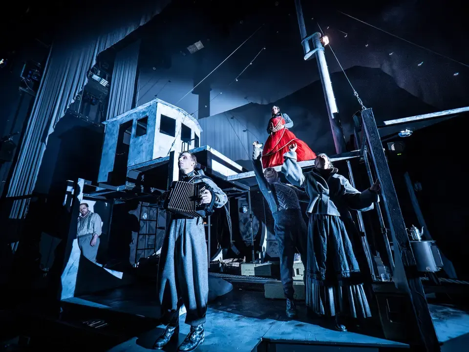 Bühnenfoto. Die Bühne erinnert an ein Schiff, die Stimmung ist kühl. Im Mittelpunkt steht eine Frau in einem roten Kleid. Weitere Figuren sind um sie herum gruppiert – einer spielt Akkordeon.